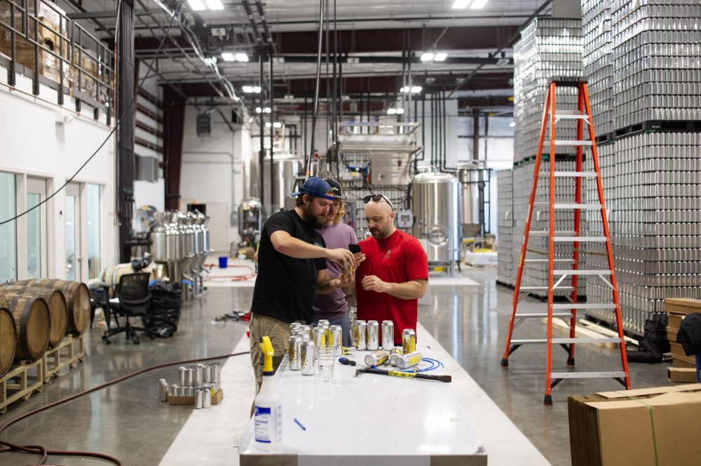 From left to right, Flying Machine head brewer Carl Cross, quality assurance manager Dean Moore, and Cask Brewing Systems rep Tim Warren inspect the label of a Vimana Kölsch can fresh out of the brewery's new canning system. (Port City Daily photo/Mark Darrough)